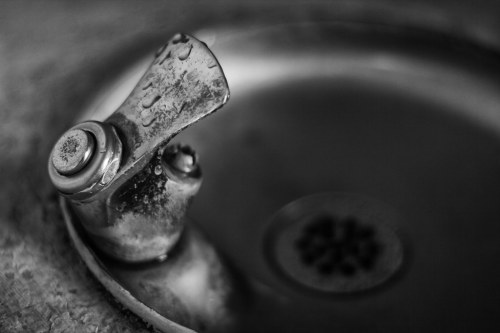 Close-up of a weathered drinking fountain spout with water droplets, set against a blurred background.