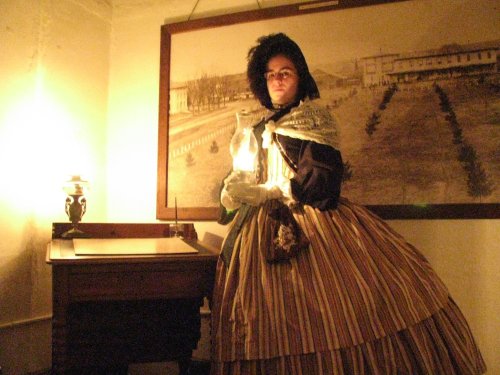 A woman in a historical dress holds a lantern, standing beside a desk in a dimly lit room with a vintage backdrop.