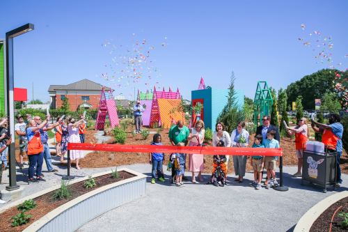 A group of people celebrates the opening of a colorful garden, cutting a ribbon as confetti falls around them.