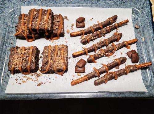 A tray of chocolate treats: rectangular bars with caramel drizzle and pretzel sticks coated in chocolate and crushed toppings.