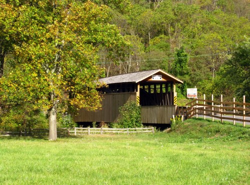 A wooden covered bridge surrounded by greenery and trees, with a grassy area in the foreground.