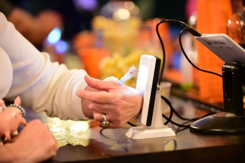 A hand holding a small object near a light source on a bar counter, with colorful drinks in the background.
