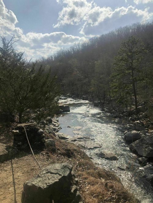 A serene river flows through a rocky landscape, surrounded by trees and a cloudy sky.