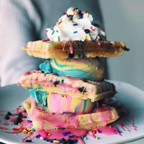 A stack of colorful ice cream between waffles, topped with whipped cream and sprinkles on a white plate.