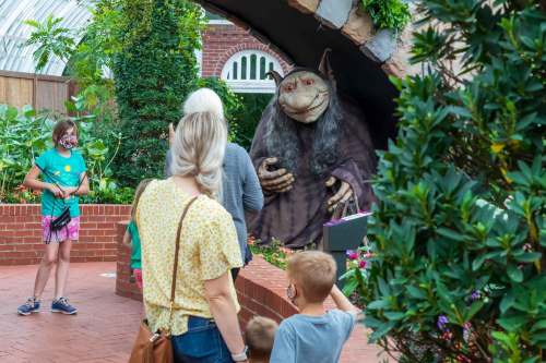 A whimsical troll statue greets visitors in a lush garden setting, with children and adults exploring nearby.