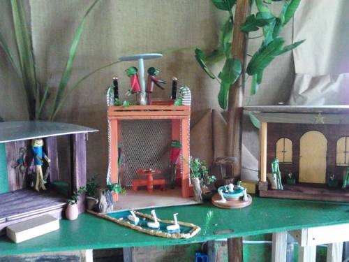 Colorful miniature structures with figures, plants, and a small pond, set against a natural backdrop.
