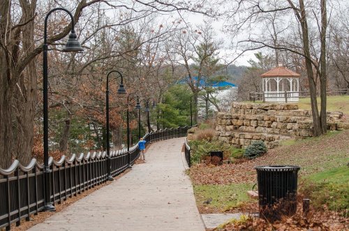A winding path through a park with a gazebo, stone wall, and trees, leading to a lake in the background.