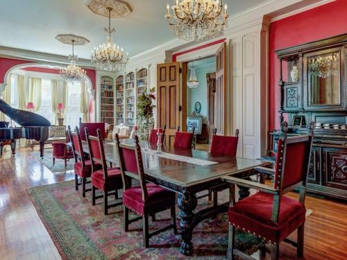Elegant dining room with a large wooden table, red velvet chairs, chandeliers, and a grand piano in a classic setting.