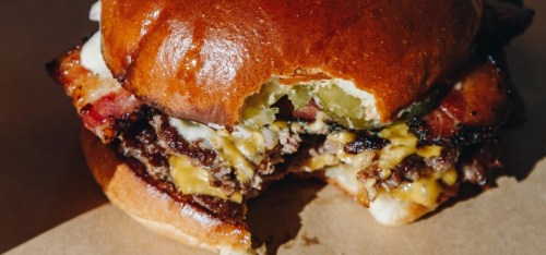 A close-up of a half-eaten burger with bacon, cheese, pickles, and a glossy bun on a brown paper surface.