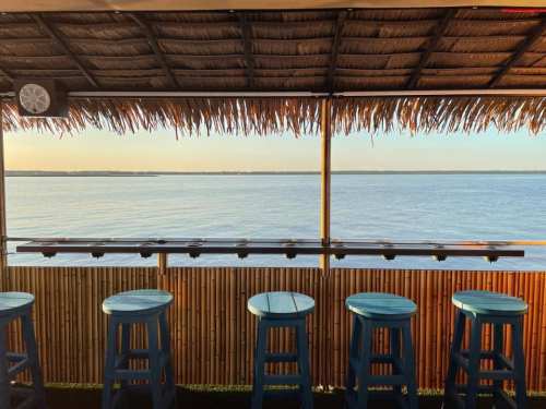 A beachside bar with blue stools overlooking a calm ocean at sunset, framed by a thatched roof.