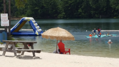 A sandy beach scene with a red chair under a straw umbrella, inflatable slide in the water, and people swimming nearby.