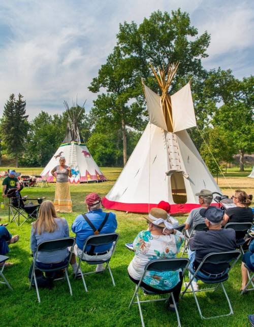 A speaker presents in front of traditional tipis, with an audience seated on chairs in a grassy area.
