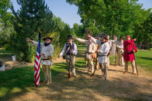 A group of historical reenactors in period clothing, holding rifles and an American flag, posing outdoors in a park.