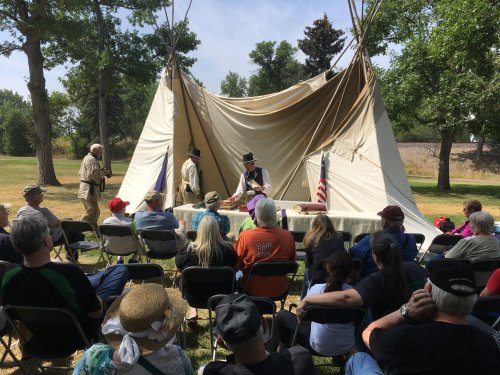 A historical reenactment with people in period costumes gathered around a tent, engaging an audience in a park setting.