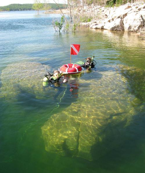 Two divers in a lake near a red and white dive flag, with a rocky underwater area visible.