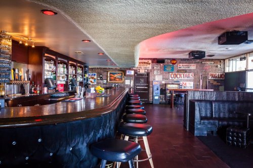 A stylish bar interior with a curved counter, stools, shelves of drinks, and a cozy seating area.