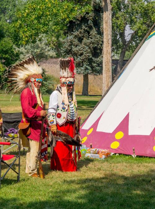 Two individuals in traditional Native American attire stand near a colorful teepee in a grassy area.