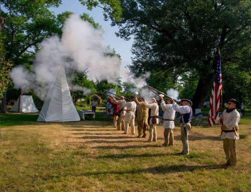 A group of reenactors in historical attire fires muskets, with smoke rising and a teepee in the background.
