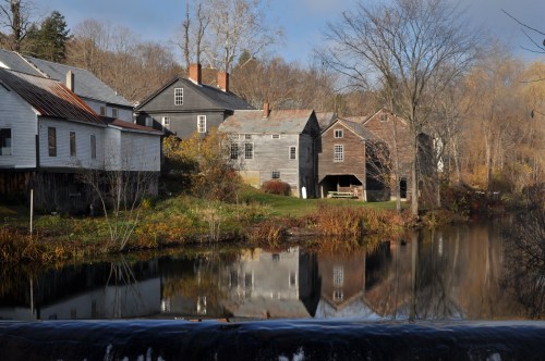 A serene scene of old houses by a calm river, reflecting autumn colors and trees in the background.