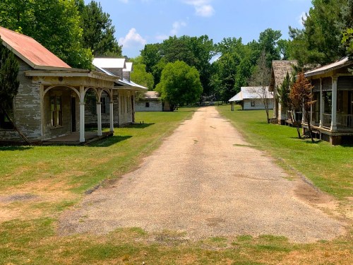 A gravel road lined with old buildings and greenery under a blue sky.
