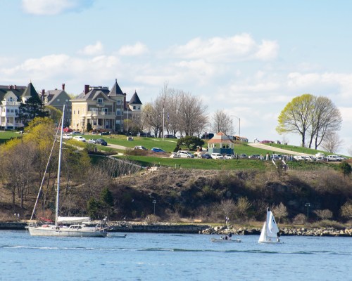 A sailboat and a small boat on a calm waterway, with a green hillside and buildings in the background under a blue sky.