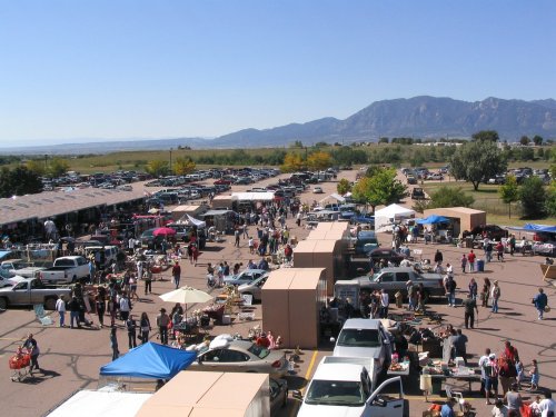 A bustling outdoor market with vendors, shoppers, and mountains in the background on a clear day.