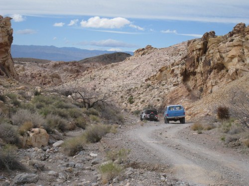A blue vehicle drives along a rocky dirt road in a desert landscape with mountains in the background.