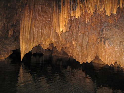 Stalactites and stalagmites in a dark cave, reflecting in still water below.