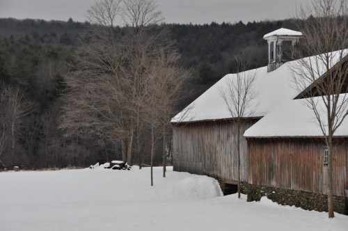 A snowy landscape featuring a rustic barn and bare trees, with a forested hillside in the background.
