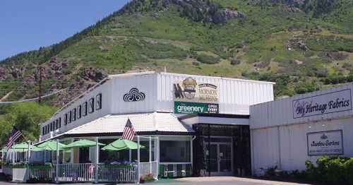 A storefront with a white exterior and green accents, set against a mountainous backdrop, featuring outdoor seating and flags.