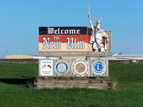 Welcome sign for New Ulm featuring a warrior figure and local organization logos against a clear blue sky.