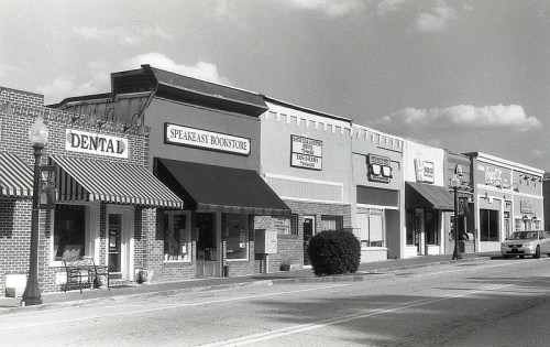 Black and white photo of a street lined with storefronts, including a bookstore and dental office, under a cloudy sky.