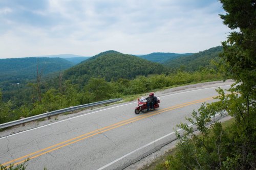 A motorcyclist rides along a winding road with lush green hills in the background under a cloudy sky.