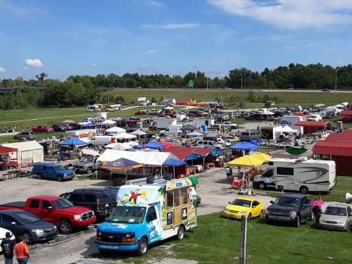 A bustling outdoor market with colorful tents, food trucks, and parked cars under a clear blue sky.