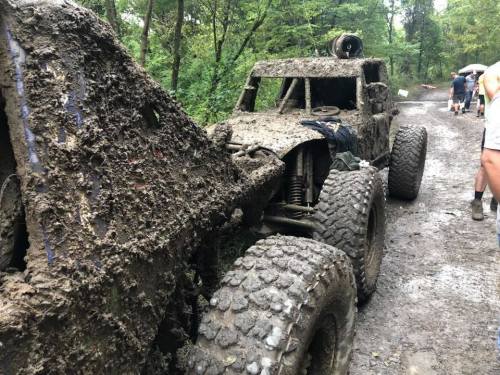 A muddy off-road vehicle parked on a dirt trail surrounded by trees, showcasing its rugged tires and dirty exterior.