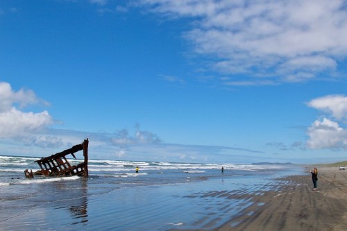 A rusted shipwreck on a sandy beach, with waves crashing and a few people walking along the shore under a blue sky.