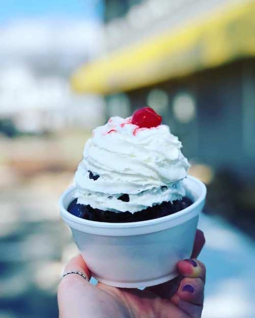 A hand holding a cup of chocolate dessert topped with whipped cream and a cherry.