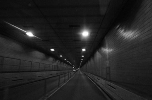 A dimly lit tunnel with a long, straight road leading into the distance, illuminated by overhead lights.