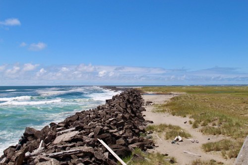 Rocky jetty extending into the ocean, with waves crashing and grassy shorelines under a clear blue sky.