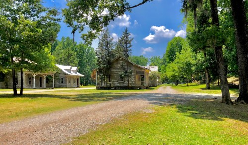 A scenic view of a gravel path leading to rustic houses surrounded by lush green trees and a blue sky.