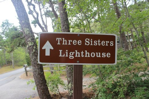 Brown sign pointing to the Three Sisters Lighthouse, surrounded by trees and greenery.