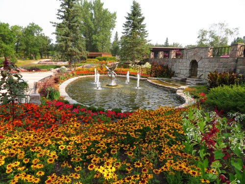 A vibrant garden with colorful flowers surrounding a pond and fountain, framed by trees and stone pathways.
