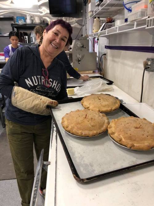 A smiling woman in an oven mitt holds a tray with three freshly baked pies in a kitchen setting.