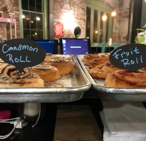 A tray of freshly baked cinnamon and fruit rolls, each labeled on small chalkboard signs.