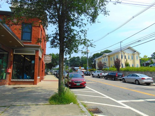 A street view featuring a brick building, parked cars, and a tree along a quiet road on a sunny day.