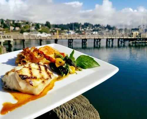 Grilled fish dish on a plate with a scenic waterfront view and boats in the background.