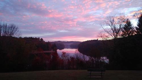 A serene lake at sunrise, surrounded by trees and colorful clouds reflecting on the water. A bench is in the foreground.