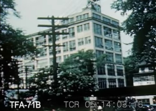 A large, multi-story building surrounded by trees and power lines, with people visible in the foreground.