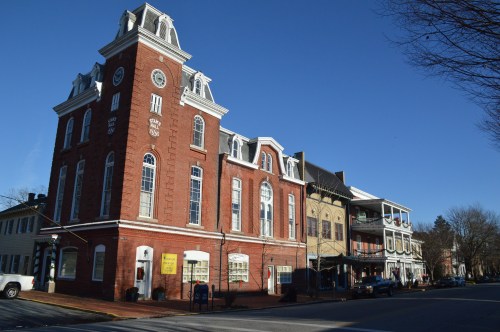 Historic brick building with clock tower beside a row of shops and a clear blue sky.