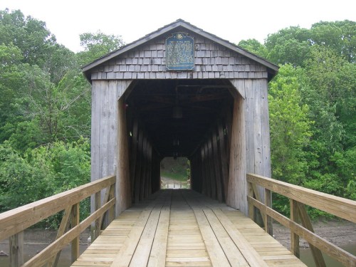 A wooden covered bridge with a plaque, surrounded by lush green trees and a wooden walkway leading inside.